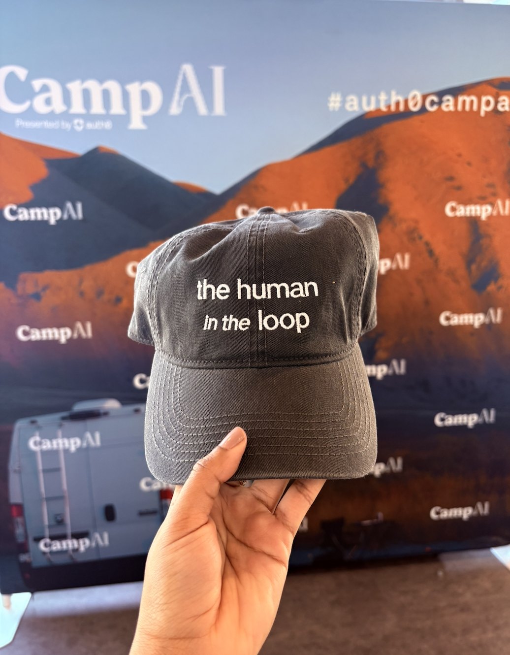 A hat that says the human in the loop in front of a Camp AI backdrop